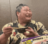 a man laughs while holding a bowl of food with chopsticks in front of a sign that says ' japan ' on it