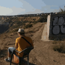 a man in a yellow shirt sits in a chair in front of a brick wall with graffiti on it that says 00