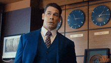 a man in a suit and tie stands in front of a wall of clocks including one for new york