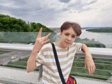 a young man in a striped shirt stands on a bridge giving a peace sign