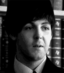 a black and white photo of a man in a suit and tie standing in front of a bookshelf .