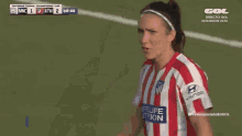 a woman in a red and white striped shirt stands on a soccer field with a score of 1 to 2