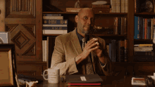 a man sits at a desk in front of a bookshelf with a mug that says i 'm a scientist on it