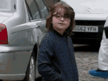 a young boy wearing glasses is standing in front of a silver car with a license plate that says sg62 yfh