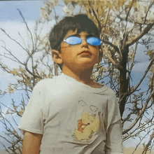 a young boy wearing sunglasses and a white shirt stands in front of trees