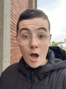 a young man wearing glasses makes a surprised face in front of a brick wall