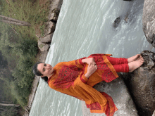 a woman in a red and orange dress sits on a rock near a river