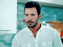 a man with a beard wearing a white shirt is standing in a kitchen