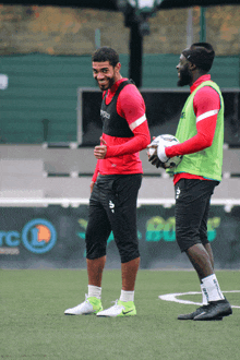 two soccer players on a field with one wearing a green vest that says asics on it