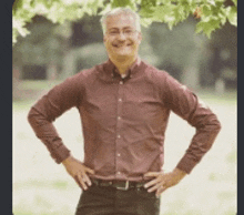 a man in a red shirt is standing with his hands on his hips in a park .