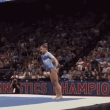 a woman in a blue leotard is doing a trick on a mat in front of a sign that says gymnastics champion