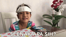 a little girl with a bandage on her head is sitting at a table with the words seenak apa sih written on it