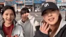 three young women are standing next to each other on a city street . one of the women is wearing a ny hat .