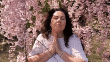 a woman is sitting under a cherry blossom tree with her eyes closed and her hands folded in prayer .