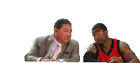 a man in a suit sits next to a man in a red shirt