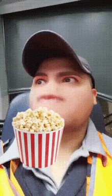 a man wearing a hat and a yellow vest is holding a striped bucket of popcorn in front of his face