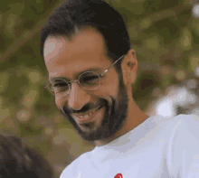 a man with glasses and a beard is smiling and wearing a white shirt with a red heart on it