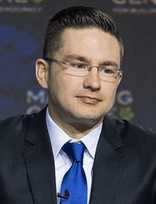 a man wearing glasses and a blue tie stands in front of a microphone .