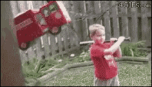 a boy in a red shirt is swinging a bat at a toy truck that is flying through the air .