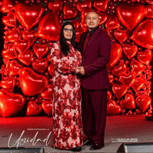 a man in a purple suit and a woman in a red dress are standing in front of a wall of heart shaped balloons