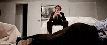 a man in a suit and tie is sitting on a couch next to a dead body .