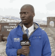 a man in a blue nbc montana jacket holds a microphone in front of a sign that says leaving yellowstone national park