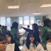 a group of students are sitting at tables in a classroom and a girl is holding a piece of paper .