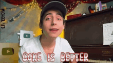 a young man wearing a hat and a white shirt is standing in front of a piano and says coke is better .