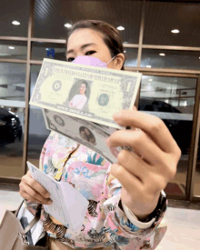 a woman wearing a pink mask is holding up a dollar bill with a picture of a woman on it