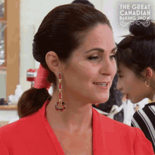 a woman wearing earrings and a red shirt is standing in front of a sign that says the great canadian baking show