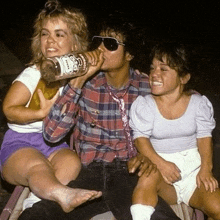 a man is drinking from a bottle while two women sit next to him on a chair .