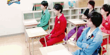 a group of young men sit at desks in a classroom with a channel logo on the wall behind them