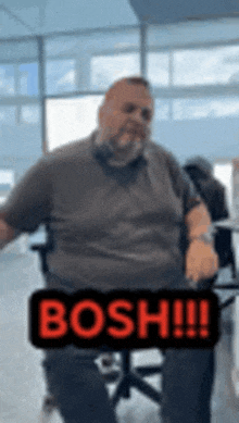 a man with a beard is sitting in an office chair with a sign that says bosh on it .