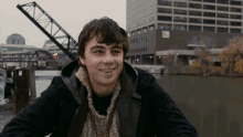 a man in a black jacket and scarf smiles in front of a building that has the letter a on it