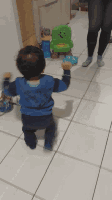 a little boy wearing a blue shirt with a shark on the back is walking towards a green toy chair