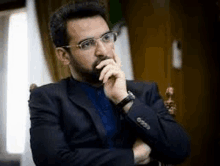 a man with a beard wearing glasses and a suit is sitting in a chair with his hand on his chin .