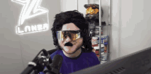 a man wearing a wig and goggles is sitting in front of a microphone in front of a computer monitor .