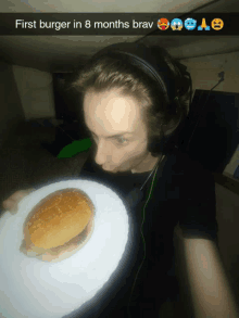 a man wearing headphones is holding a plate with a hamburger on it and a caption that says first burger in 8 months brav