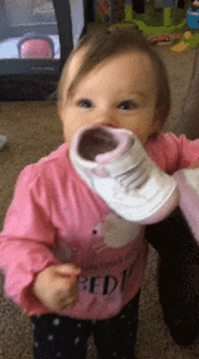 a baby wearing a pink shirt with the word red on it is holding a white shoe in her mouth