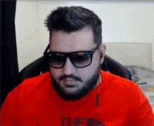 a man with a beard and sunglasses is wearing a red shirt and sitting in a chair .