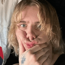 a young man with long blonde hair is making a funny face with his hand covering his mouth .