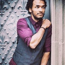 a man leaning against a wall wearing a purple shirt and a grey vest