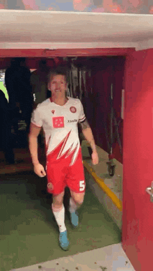 a man in a red and white soccer uniform with the number 5 on his shorts is walking out of a tunnel .