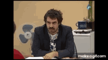 a man in a suit sits at a desk in front of a fan and a make a gif.com logo