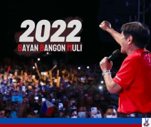 a man in a red shirt stands in front of a crowd with a fist in the air and the year 2022