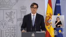 a man in a suit and tie stands at a podium in front of spanish flags and a sign language interpreter