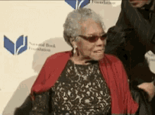 an elderly woman wearing sunglasses and a red scarf is sitting in front of a wall that says national book foundation