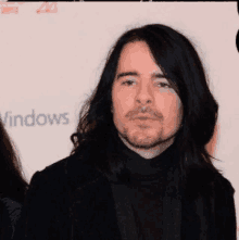 a man with long black hair and a beard stands in front of a windows sign