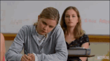 a girl is sitting at a desk in a classroom writing on a piece of paper while another girl looks on .