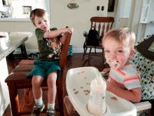 a boy in a tiger shirt sits on a chair next to a girl in a high chair drinking from a bottle